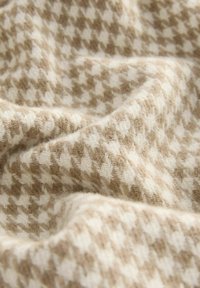 Beżowa i kremowa tkanina w wzór houndstooth, wykonana z miękkiego materiału dzianinowego o teksturowanym wykończeniu, z delikatnymi fałdami i zwisami.