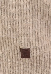 Beige gestrickter Pullover mit einem gerippten Muster, versehen mit einem quadratischen braunen Lederetikett in der unteren linken Ecke, auf dem der Markenname steht.