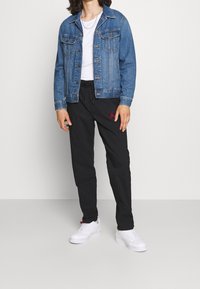 Blaue Jeansjacke mit Metallknöpfen, kombiniert mit schwarzen Jogginghosen mit rotem Logo und weißen Sneakers mit strukturiertem Finish.
