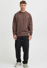 Brun fleece sweatshirt med rund hals og hvid "A.R.C." logo på venstre bryst, sammen med sorte bukser og lyse sneakers.
