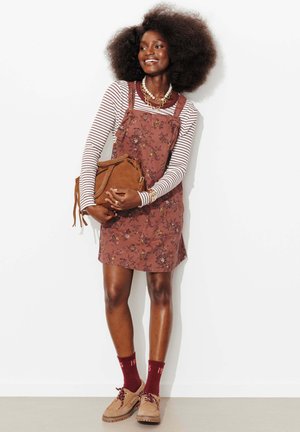 Femme souriante avec une grande afro portant un t-shirt rayé à manches longues sous une robe fleurie, tenant un sac marron, avec des chaussettes bordeaux et des chaussures beige.