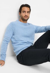 Helles blaues, gestricktes Sweatshirt mit einem strukturierten Muster und Rundhalsausschnitt, kombiniert mit schwarzen, enganliegenden Hosen. Der Hintergrund ist neutral.