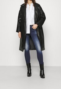 Svart läderlångjacka med bred krage, vit skjorta, mörkblå jeans och svarta klackstövlar. Slät textur, stilren design.