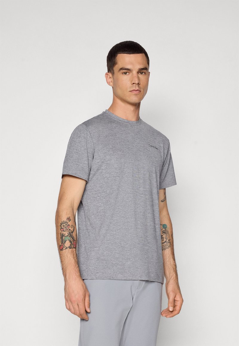 T-shirt grigia a maniche corte in tessuto leggero con colletto girocollo e logo discreto sul petto, abbinata a pantaloni grigi chiari.