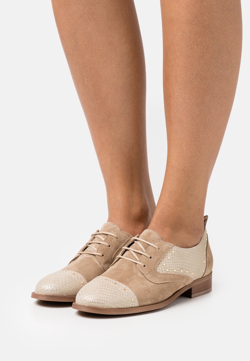 Chaussures lacées en daim marron clair avec des panneaux beiges texturés sur l'avant et les côtés, portées pieds nus sur fond blanc.