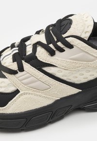 Klobige Sneaker mit einem strukturierten beige Obermaterial, schwarzen Akzenten und perforierten Paneelen. Mit synthetischen Schnürsenkeln und einer Gummisohle für Traktion.