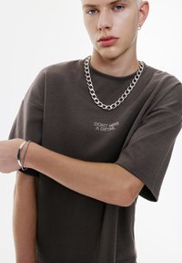 Brun oversized t-shirt med korte ærmer og broderet tekst "GÅ IKKE GLIP AF EN DETALJE." Sølv kæde halskæde og armbånd, glat stofstruktur.