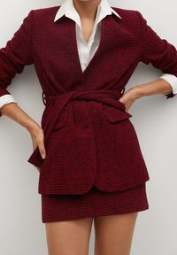 Czerwony żakiet z tkaniny tweedowej z paskiem w talii, zestawiony z białą koszulą z kołnierzykiem i pasującą spódnicą, z fakturowanym wzorem.