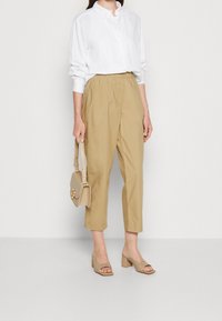 Donna che indossa pantaloni cropped beige larghi, camicia bianca a maniche lunghe con bottoni, sandali con tacchi beige e porta una borsa beige.