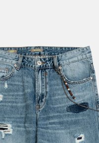 Jeans de mezclilla con lavado azul claro, patrones desgastados, detalles metálicos y cadena decorativa colgando del bolsillo delantero.
