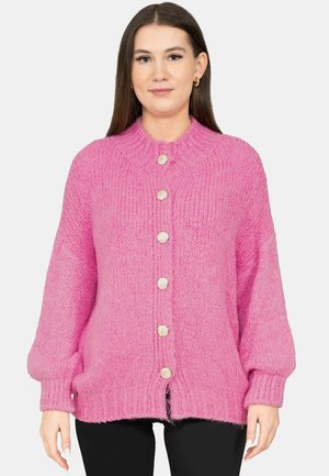 Femme portant un cardigan tricoté épais rose vif avec six gros boutons, debout devant un fond clair uni.