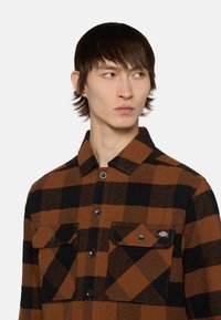 Brun og sort ternet flannel skjorte med lange ærmer, knaplukning foran, to brystlommer og mærke på venstre lomme.