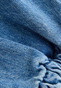 Denim tkanina svijetloplavog do srednje plavog tona, s teksturiranim tkanjem i naglašenim detaljima, prikazujući mekanu, pomalo ugužvanu površinu.