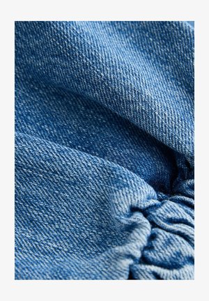 Denimkangas, millel on hele kuni keskmine sinine tooni, tekstuurse kanga ja kogutud detailidega, mis näitab pehmet, veidi kortsus pinda.