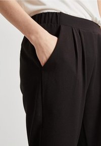 La mano è appoggiata all'interno della tasca anteriore di pantaloni neri con vita elastica, abbinati a un top bianco.