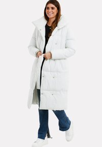 Hvid pufferjakke med høj krave og trykknapper, med en quiltet design. Båret over en sort top og parret med denim og sneakers.