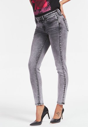 Kvinde iført gråslidte skinny jeans parret med sorte høje hæle, hånd hvilende på hoften, synlig tatovering på venstre ankel.