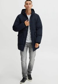 Abrigo acolchado azul marino con capucha, que cuenta con una cremallera frontal, botones a presión, dos bolsillos laterales y una textura acolchada. Usado sobre una camisa blanca.