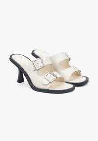 White heeled sandals with two silver buckles, open-toe design, and a textured black outsole. Elegant curved heel and cut-out sections.