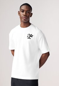 T-shirt en coton blanc à manches courtes avec un chat noir de dessin animé sur un skateboard, situé sur la partie gauche de la poitrine.