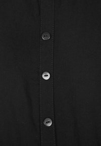 Chemise noire à boutons, présentant six boutons noirs ronds le long de la patte de boutonnage, avec une texture en coton lisse et des détails de couture visibles.