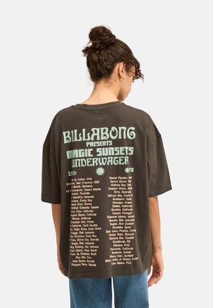 Osoba o ciemnych włosach związanych w kok, ubrana w brązowy, oversize’owy t-shirt Billabong z jasnoniebieskim napisem na plecach wymieniającym globalne lokalizacje surfingowe.