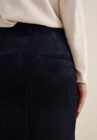 Jupe en velours côtelé navy avec une texture à côtes verticales, dotée d'une taille haute et de deux poches arrière discrètes, partiellement couverte par un pull beige.