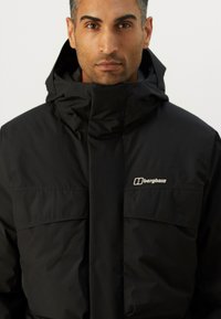 Mand med kort gråt hår iført en sort Berghaus-hættetrøje med to brystlommer, set forfra mod en ensartet lys baggrund.