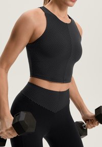 Čierny krátky top s vertikálnymi bielymi pruhmi, v páre s čiernymi legínami s vysokým pásom. Modelka drží šesťuholníkové činky v oboch rukách.