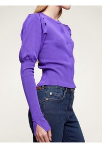 Lilafarbenes Rippstrickpullover mit Puffärmeln, taillierter Silhouette und Daumenlöchern, kombiniert mit dunklen Jeans.