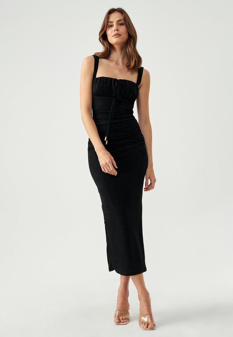 Black fitted dress with a ruched design, square neckline, and thick straps, featuring a side split and smooth texture. Styled with clear heels.