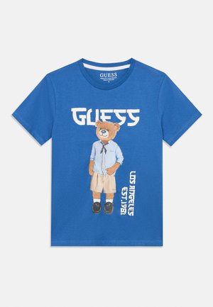 T-shirt bleu à manches courtes avec un ours portant une chemise, un short et des chaussures, texte « GUESS » au-dessus, et texte vertical « Los Angeles Est. 1981 » sur le côté.