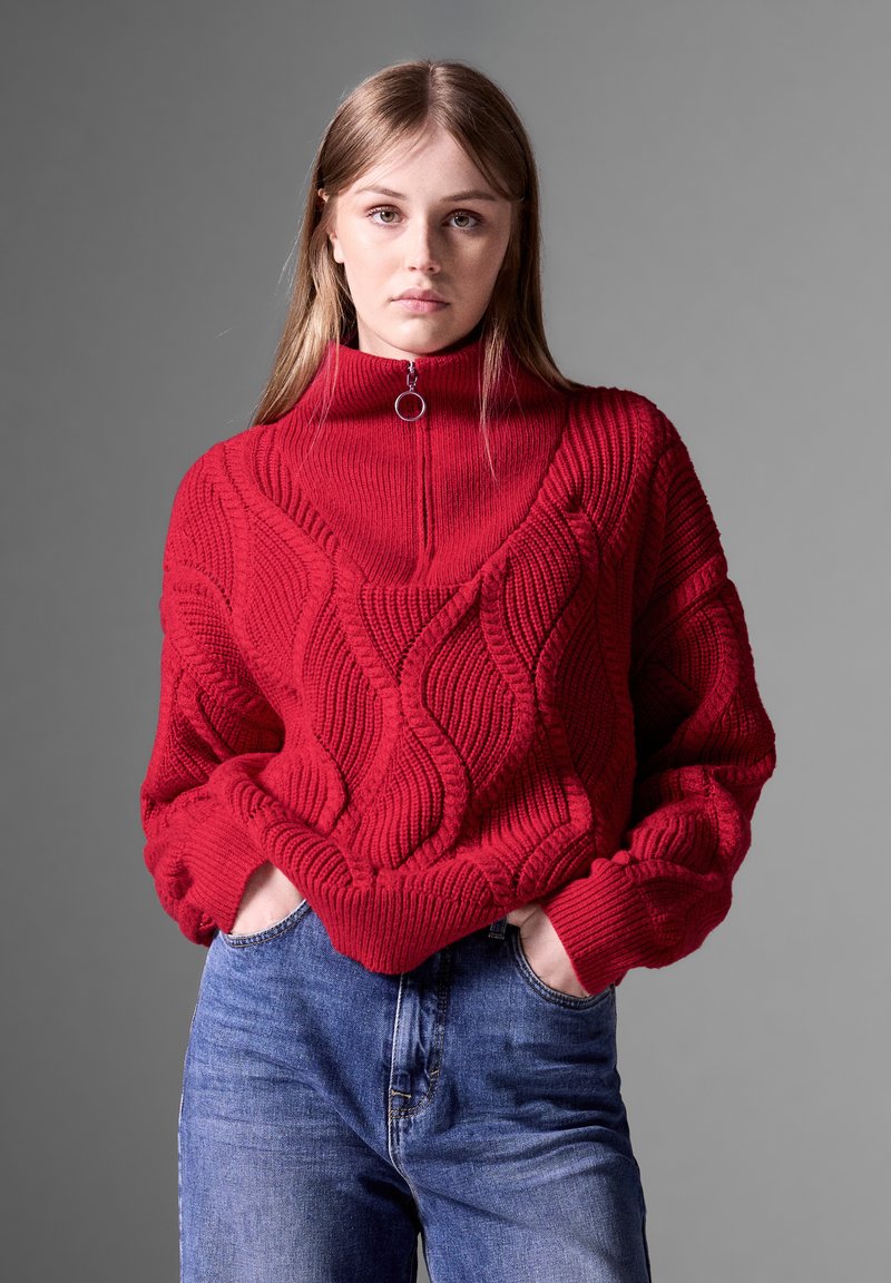 Roter Strickpullover mit hohem Kragen, der strukturierte Wellenmuster aufweist. Kombiniert mit blauen Jeans im lässigen Schnitt.