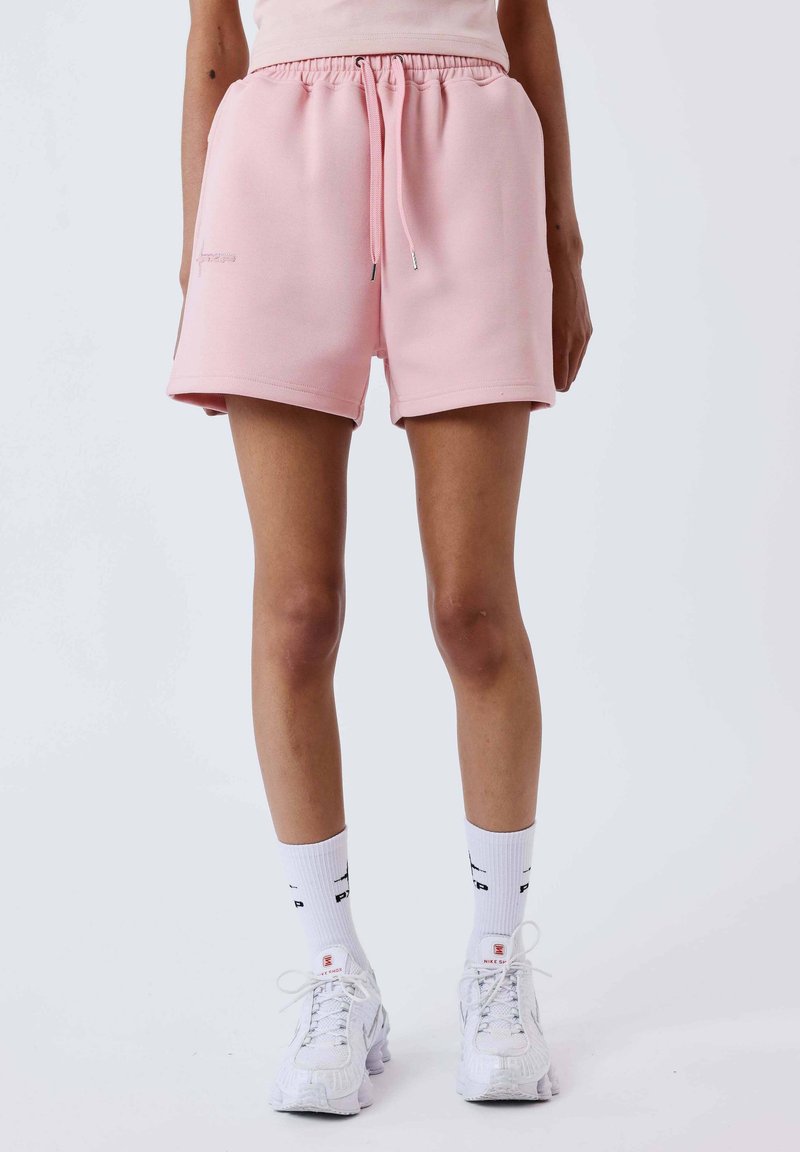 Personne portant un short à cordon de serrage rose clair, des chaussettes de sport blanches mi-mollet avec du texte noir, et des baskets blanches, debout sur un fond uni.
