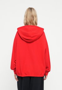 Felpa rossa oversize con cappuccio regolabile e fondo a costine. Realizzata in tessuto morbido, presenta una superficie liscia e un design semplice.