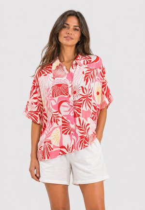 Vrouw met lang haar die een roze en rood patroon korte mouwen blouse met knopen en witte korte broek draagt, staand voor een effen achtergrond.