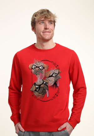 Joven con una sudadera roja con un gráfico de tres carlinos juguetones que llevan gafas y sacan la lengua.