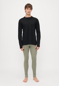 Crna majica dugih rukava i maslinasto zelene uske hlače, obje izrađene od glatkog materijala. Model stoji bos, prikazujući cijeli outfit.