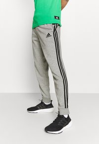 Pantalones de chándal grises en un diseño ajustado con rayas laterales negras, cuentan con una cinturilla elástica y puños en los tobillos, combinados con zapatillas deportivas negras.