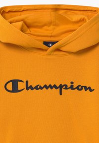 Felpa arancione in morbido tessuto con cappuccio regolabile e un grande logo "Champion" nero sul petto.