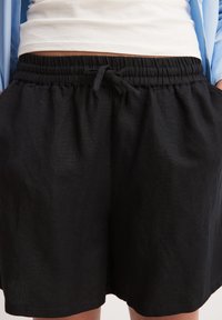 Pantaloni corti neri realizzati in tessuto testurizzato con vita elastica, dotati di un nodo frontale e tasche laterali, progettati per comfort e facilità di indossamento.