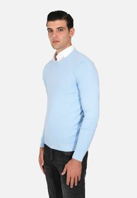 Maglione lavorato a maglia blu chiaro con collo a giro e polsini a costine, indossato sopra una camicia bianca con colletto, abbinato a jeans scuri.