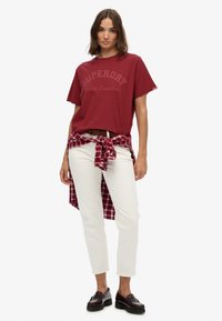 Rode katoenen T-shirt met "Superdry" logo, witte broek en een geknoopte geruite overhemd. Zwarte instappers en een bruine gevlochten riem maken de uitstraling compleet.