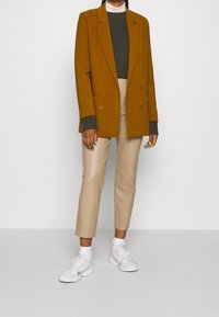 Blazer marron croisé, pull vert texturé, pantalon beige slim, baskets blanches, présenté sur un fond uni.