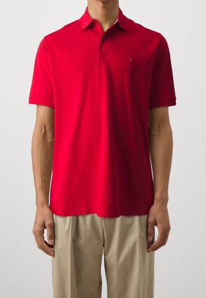 Röd polo skjorta i slät tyg med krage, korta ärmar och en liten logga på vänster bröst. Kombinerad med khakifärgade byxor.