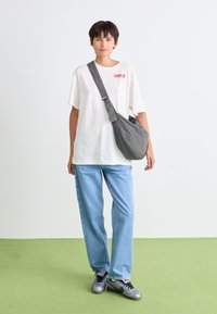 Bílý tričko s červeným logem, modré džíny a šedobéžová crossbody taška. Šedé tenisky s černými tkaničkami dotvářejí outfit.