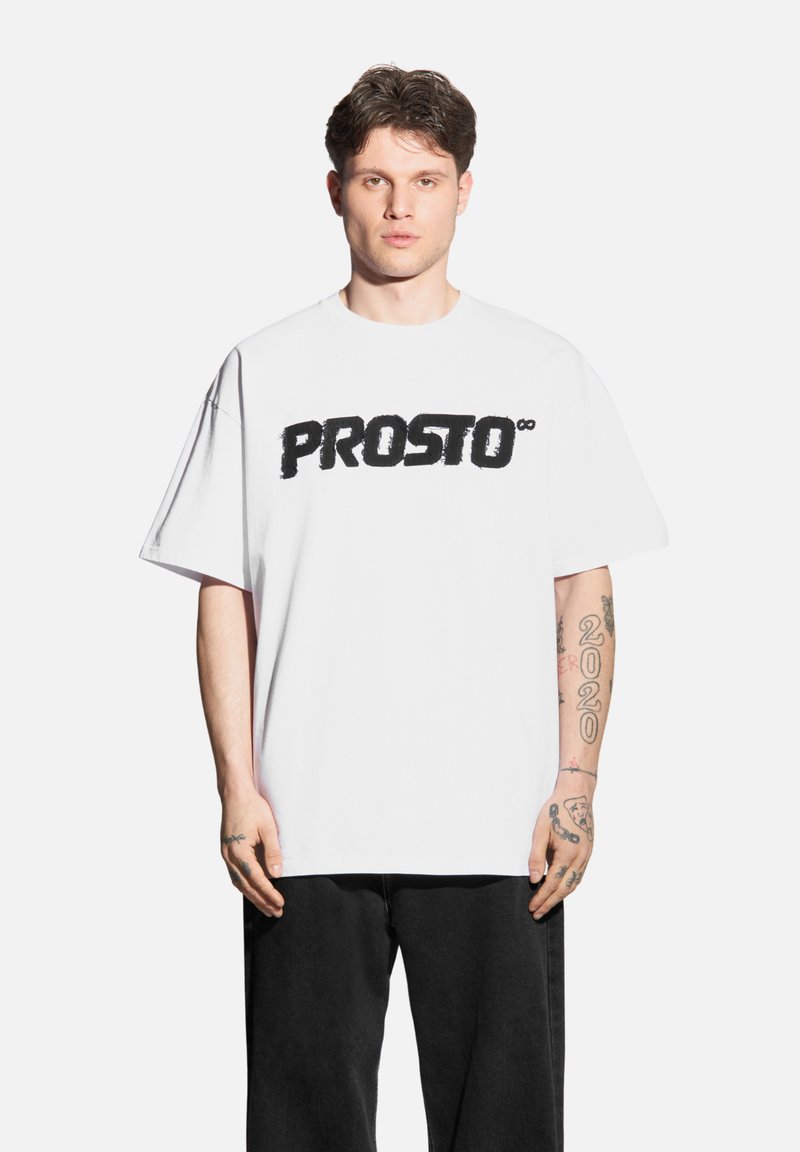 Biała bawełniana koszulka z krótkim rękawem z napisem "PROSTO" w wyraźnej czarnej czcionce. Luźny krój, noszona z czarnymi spodniami, z widocznymi tatuażami.