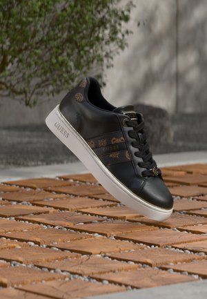 Basket Guess noire avec des accents marron à motifs et une semelle blanche, flottant au-dessus de carreaux en bois en extérieur avec un fond flou de verdure et de mur.