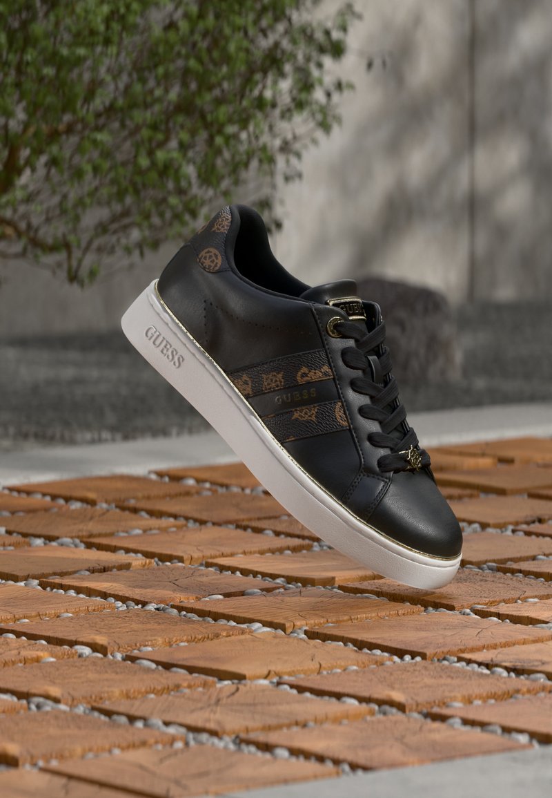 Basket Guess noire avec des accents marron à motifs et une semelle blanche, flottant au-dessus de carreaux en bois en extérieur avec un fond flou de verdure et de mur.