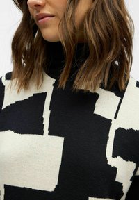 Schwarzer und cremefarbener Strickpullover mit Rollkragen, der abstrakte Blockmuster aufweist, mit einer weichen Textur und tailliertem Design.